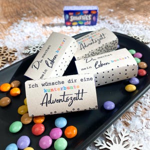 Könnte beinhalten: Vier kleine braune Schachteln mit weißen Punkten und buntem Text. Die Schachteln haben den Text "Du machst mein Leben bunter!" und "Ich wünsche dir eine kunterbunte Adventszeit!" aufgedruckt. Die Schachteln sind von bunten Süßigkeiten umgeben.