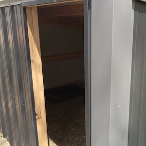 May include: A grey metal shed with a sliding door. The interior reveals wooden beams and a floor covered in hay. The shed is outdoors, with a glimpse of the sky and grass visible.