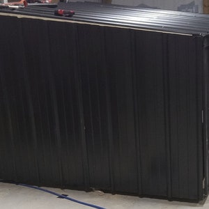 May include: A black metal shed with a sloping roof. The shed features vertical corrugated siding and is under construction. Tools are visible on the roof. The shed is likely used for storage.