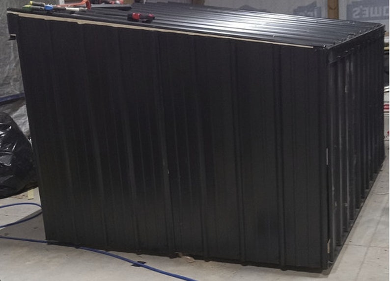 May include: A black metal shed with a sloping roof. The shed features vertical corrugated siding and is under construction. Tools are visible on the roof. The shed is likely used for storage.