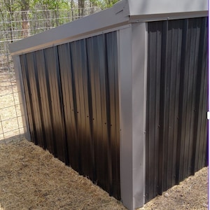 May include: A black metal shed with a grey roof and trim. The shed features vertical corrugated panels and is set on straw. The structure is outdoors, with a wire fence and trees in the background.