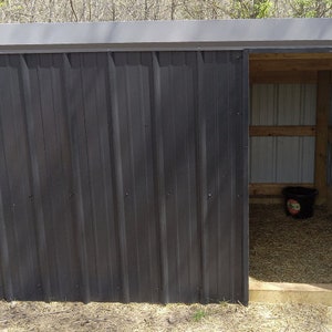 May include: A small, dark grey metal shed with a sliding door. The shed has a corrugated metal exterior and a wooden interior. A black bucket sits inside the shed on a bed of hay. The shed is outdoors, with trees in the background.