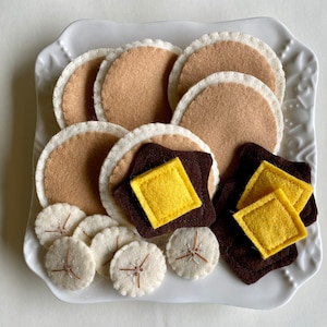 May include: A white plate with felt food items, including brown pancakes, yellow butter pats, and brown chocolate squares. There are also white felt banana slices on the plate.