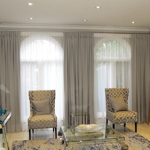 May include: A living room with three arched windows, each with a pair of curtains. The curtains are a light grey colour and are drawn back to reveal sheer white curtains behind them. Two chairs with patterned upholstery are positioned in front of the windows, facing each other. A glass coffee table sits between the chairs.