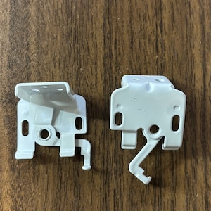 May include: Two white metal brackets with a lever mechanism. The brackets are designed for mounting window blinds.