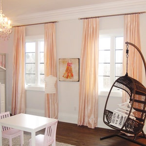 May include: A light pink and white bedroom with a white table and two pink chairs. There are pink curtains on the windows and a hanging wicker chair in the corner. There is a painting of a dress on the wall.