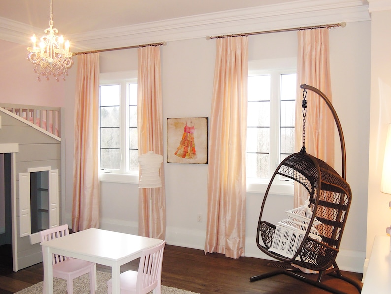 May include: A light pink and white bedroom with a white table and two pink chairs. There are pink curtains on the windows and a hanging wicker chair in the corner. There is a painting of a dress on the wall.