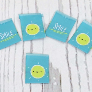 May include: Five small, rectangular fabric tags with a light blue background. Each tag features a smiling sun graphic in yellow and white, with the word "SMILE" in white. The tags are arranged on a white wooden surface.
