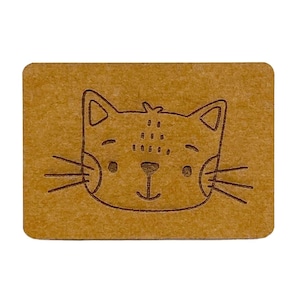 Op de afbeelding: Een bruin papieren label met een schattig cartoon kattengezicht erop getekend. De kat heeft een zwarte omtrek en een witte neus en ogen.