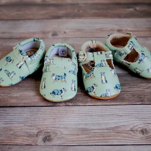 May include: Four pairs of baby shoes with a blue and white dog print on a light green background. Two pairs are slip-on style and two pairs are t-strap style.