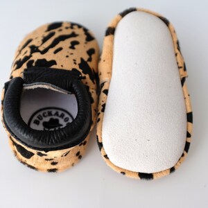 May include: A pair of baby moccasins with a black and tan cowhide print. The moccasins have a black leather cuff with the word "BUCKARO" in white lettering and a small cowboy hat logo.