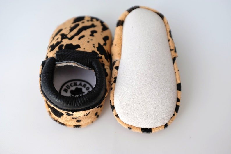 May include: A pair of baby moccasins with a black and tan cowhide print. The moccasins have a black leather cuff with the word "BUCKARO" in white lettering and a small cowboy hat logo.