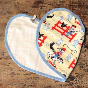 Puede incluir: Un babero de algod&oacute;n blanco en forma de coraz&oacute;n, con ribete azul, con un dise&ntilde;o impreso en amarillo y blanco de vaqueros y caballos. El dise&ntilde;o incluye una valla roja, cactus y un vaquero montando un caballo.
