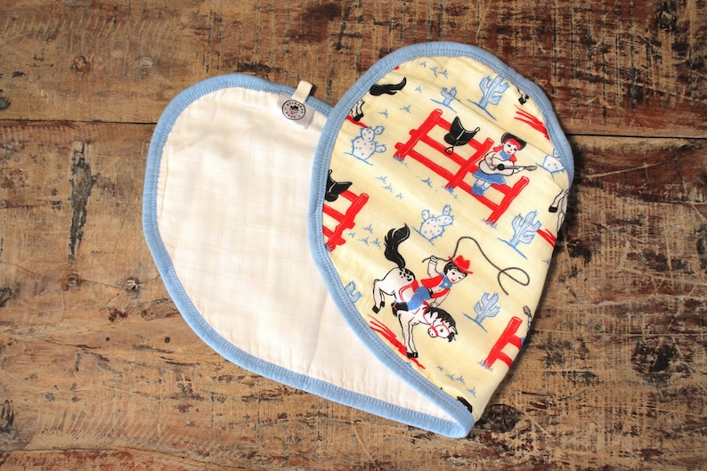 Puede incluir: Un babero de algod&oacute;n blanco en forma de coraz&oacute;n, con ribete azul, con un dise&ntilde;o impreso en amarillo y blanco de vaqueros y caballos. El dise&ntilde;o incluye una valla roja, cactus y un vaquero montando un caballo.