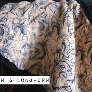 Puede incluir: Una tela beige con un estampado en blanco y negro de cabezas de bisontes y longhorn. La tela tiene un ribete negro alrededor de los bordes. El texto "BISON & LONGHORN" est&aacute; impreso en la tela.
