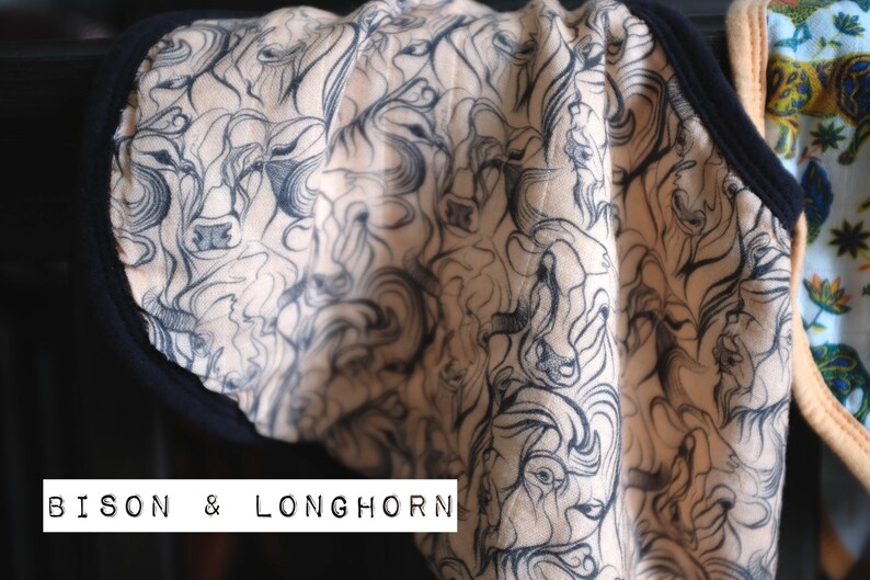 Puede incluir: Una tela beige con un estampado en blanco y negro de cabezas de bisontes y longhorn. La tela tiene un ribete negro alrededor de los bordes. El texto "BISON & LONGHORN" est&aacute; impreso en la tela.