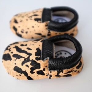 May include: A pair of baby moccasins with a black and tan animal print design. The moccasins have a black leather trim around the opening.