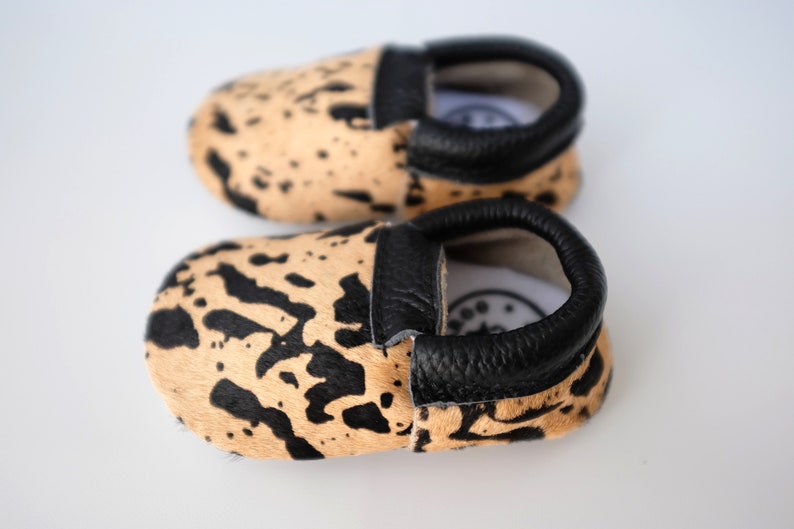 May include: A pair of baby moccasins with a black and tan animal print design. The moccasins have a black leather trim around the opening.