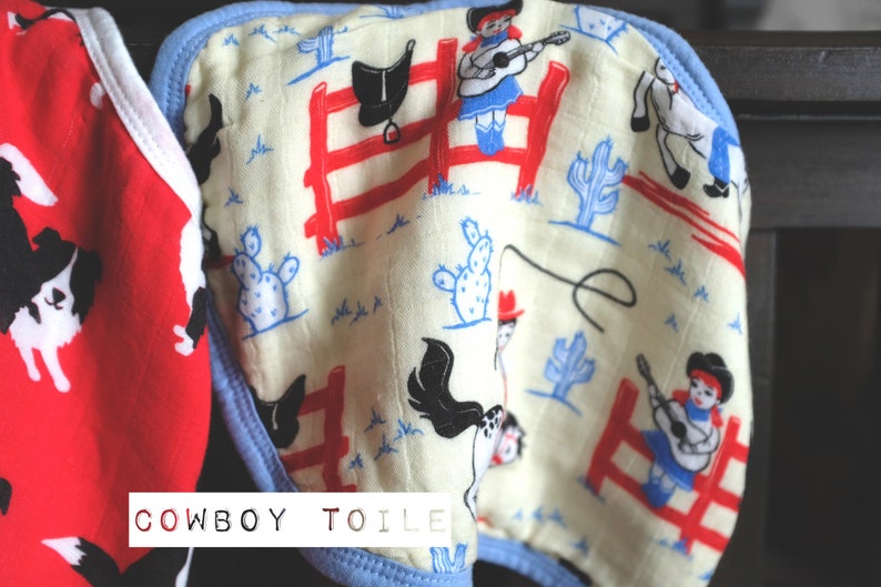 Puede incluir: Dos baberos para beb&eacute; con tem&aacute;tica de vaquero. Un babero es rojo con perros border collie negros y blancos. El otro babero es blanco con un borde azul y un patr&oacute;n de vaqueros, vaqueras, caballos, cactus y sillas de montar. El texto "COWBOY TOILE" est&aacute; impreso en la parte inferior de los baberos.