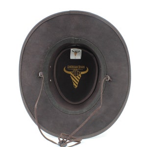 Pu&ograve; includere: Cappello da cowboy in pelle marrone con cinturino in pelle marrone e fibbia nera. Il cappello &egrave; foderato in feltro nero con un logo dorato che dice "American Bison" e una testa di bisonte stilizzata con un motivo a bandiera americana.