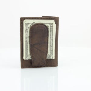 May include: A brown leather wallet with a money clip. The wallet has a fold-over design and is closed with a snap. The wallet is filled with cash.