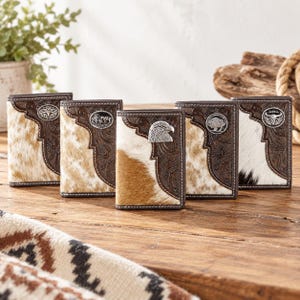 May include: Five bi-fold wallets featuring a combination of brown tooled leather with floral patterns and patches of natural hair-on hide in various shades of tan, white, and black, each accented with a silver-toned metal emblem.