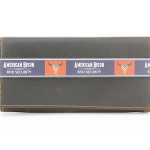 May include: A dark brown leather wallet featuring a horizontal paper band with the text AMERICAN BISON RFID SECURITY and a graphic of a bull skull with an American flag pattern. The wallet has visible stitching along the edges and a rectangular shape.