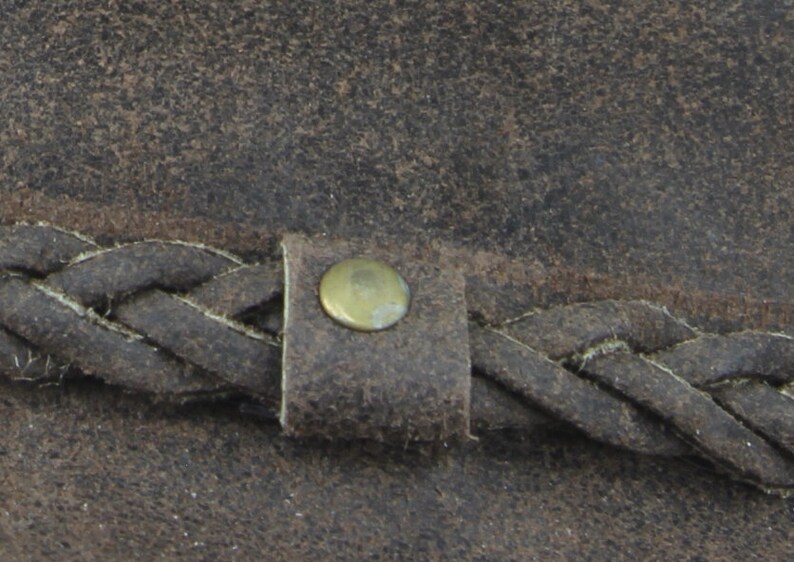 Pu&ograve; includere: Primo piano di un cappello di pelle marrone con una fascia in pelle intrecciata e un bottone in ottone.