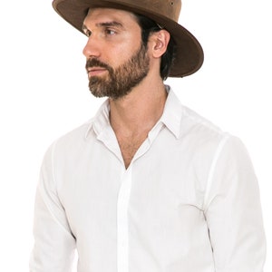 Pu&ograve; includere: Un uomo che indossa un cappello di pelle marrone a tesa larga e una camicia bianca abbottonata.