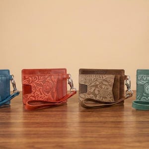 May include: Four leather card wallets in blue, red, brown, and green, each with a floral embossed design. They have wrist straps and silver-tone hardware. The wallets are displayed on a wooden surface.