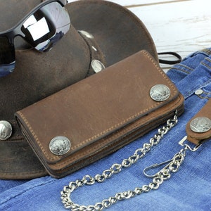 May include: A brown leather wallet with a chain and a silver concho on each flap. The wallet is lying on a pair of blue jeans.