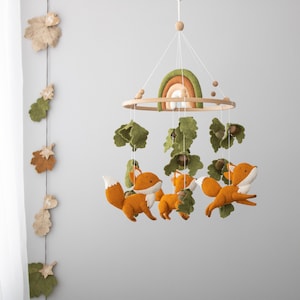May include: A baby mobile featuring orange foxes, green leaves, and a rainbow. The mobile is suspended from a wooden ring and includes wooden beads. The foxes have white bellies and tails. The leaves and acorns are felt.