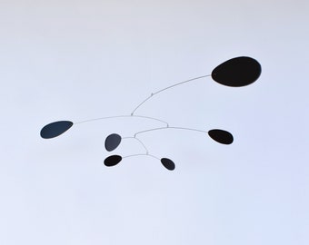 Peut inclure: Une sculpture mobile noire avec cinq pièces ovales suspendues à de fins fils. La mobile est suspendue au plafond sur un fond blanc.