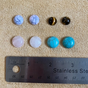 May include: Assortment of eight round cabochon gemstones, including white, pink, turquoise, and tiger's eye. The stones are arranged in two rows of four on a brown surface. A ruler is visible at the bottom of the image.