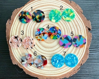 Puede incluir: Surtido de 7 cabujones redondos de resina, coloridos y con patrones, cada uno con un agujero para la elaboración de joyas. Los cabujones están dispuestos en dos filas sobre una superficie de madera.