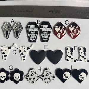 Puede incluir: Una colección de pendientes con temática de Halloween. Los diseños incluyen una máscara de Scream, ataúdes con texto, fantasmas, calaveras de vaca, corazones negros, botas con estampado de vaca, calaveras de flores, fantasmas con flores y calaveras de corazón.