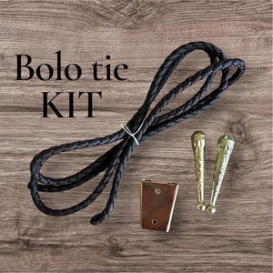 May include: A bolo tie kit with a black braided cord, a rectangular metal clasp, and two decorative gold-colored tips. The text "Bolo tie KIT" is displayed in black letters.