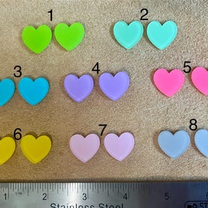 Op de afbeelding: Acht paar hartvormige oorbellen in verschillende kleuren, waaronder lichtblauw, lichtgroen, roze, geel en paars. Elk paar is genummerd van 1 tot 8.