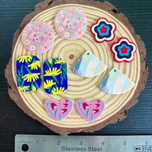 May include: A selection of colourful acrylic earrings in various shapes, including circles with bunny designs, flower shapes, triangles, and hearts. The earrings feature floral and abstract patterns in shades of pink, blue, yellow, and red. The earrings are displayed on a wooden slice.