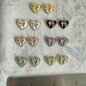 Op de afbeelding: Een verzameling hartvormige bedels in verschillende kleuren, waaronder geel, wit, zwart, roze, paars, blauwgroen en groen. Elke bedel heeft een gouden strik. Een liniaal is geplaatst voor de maatreferentie.