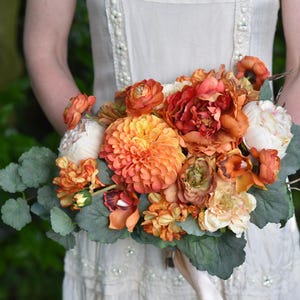 May include: A vibrant bouquet featuring orange, peach, and cream-colored flowers, including dahlias and peonies, accented with green foliage and delicate branches. The bouquet is held against a soft, off-white dress, creating a natural and elegant aesthetic.