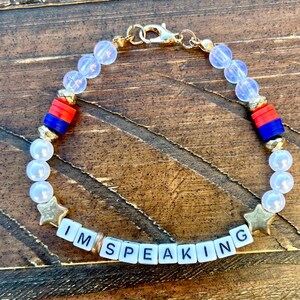 Puede incluir: Un brazalete de cuentas con cuentas blancas, estrellas doradas y cuentas de letras que deletrean "I'M SPEAKING". El brazalete también tiene cuentas rojas, azules y doradas.