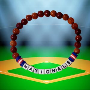 May include: A bracelet made of wooden beads and white letter beads spelling "NATIONALS". The bracelet has red, white, and blue accent beads. The background shows a baseball field.