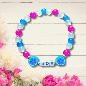 May include: A pink, blue and white beaded bracelet with a flower charm and the word "JOY" spelled out in white letters.