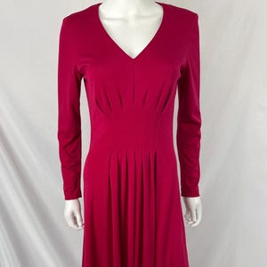 May include: A long, crimson dress with a V-neck and long sleeves. The dress features pleats at the waist and a flowing skirt. The garment is displayed on a mannequin against a white backdrop, showcasing its elegant design.