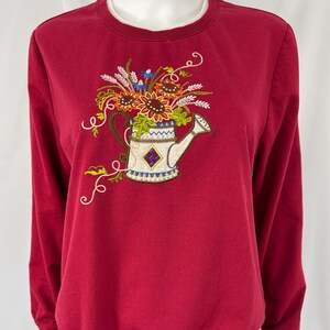 May include: A red sweatshirt featuring an embroidered design of a watering can filled with sunflowers and other flowers. The watering can is white with blue and yellow accents. The sweatshirt has long sleeves and a crew neck.