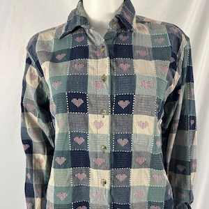 May include: A button-down shirt with a patchwork design of squares in shades of blue, green, and beige. Each square features a small, red heart pattern. The long-sleeved shirt has a collared neckline and is displayed on a mannequin.