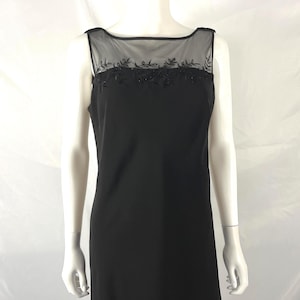 May include: A sleeveless black dress featuring a sheer mesh yoke at the neckline. The mesh panel is decorated with black floral embroidery and subtle beadwork. The dress has a straight silhouette and a wide, rounded boat neckline design.