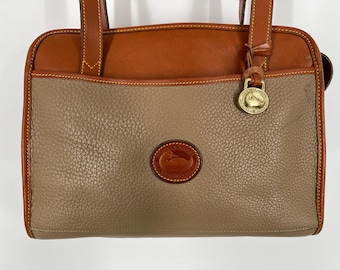 May include: A two-tone leather handbag with a tan body and brown top and trim. The bag features a gold-tone clasp and a circular logo. The bag has two handles and is designed for carrying.
