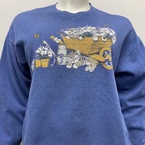 May include: A blue long-sleeved sweatshirt with a graphic of kittens in a wheelbarrow surrounded by flowers and a watering can.